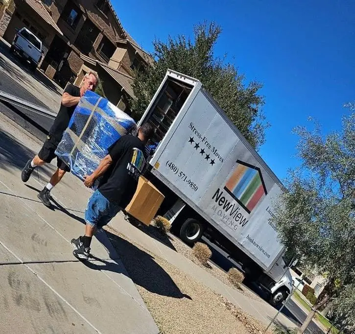 Two NewView movers carry a shrink-wrapped dresser to the company truck